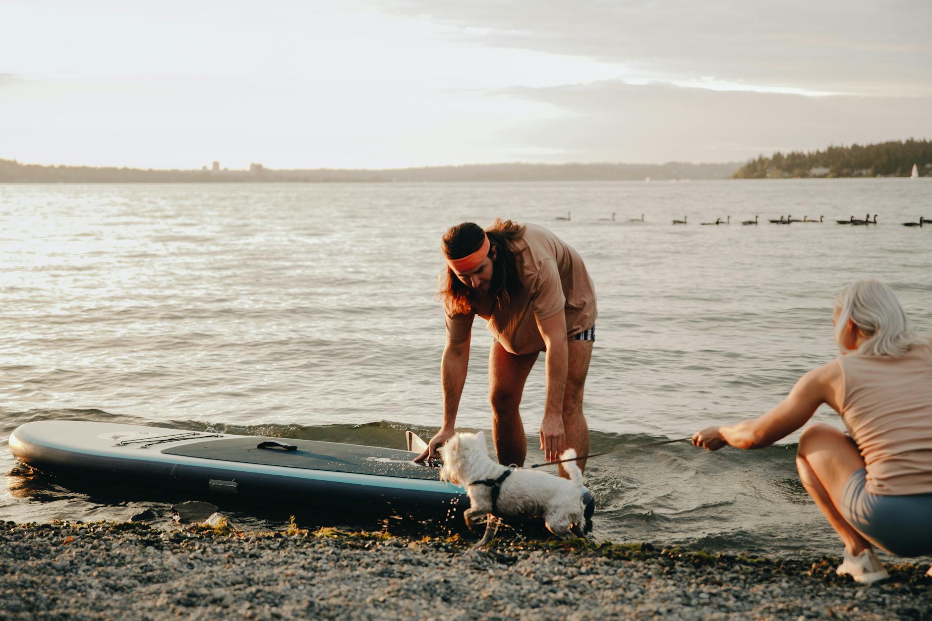 man putting a sup board into the water and woman holding a dog